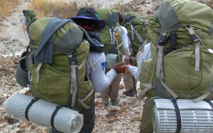 a group of students carrying backpacks descend over a rocky landscape. two of the students hold hands as they hike. 